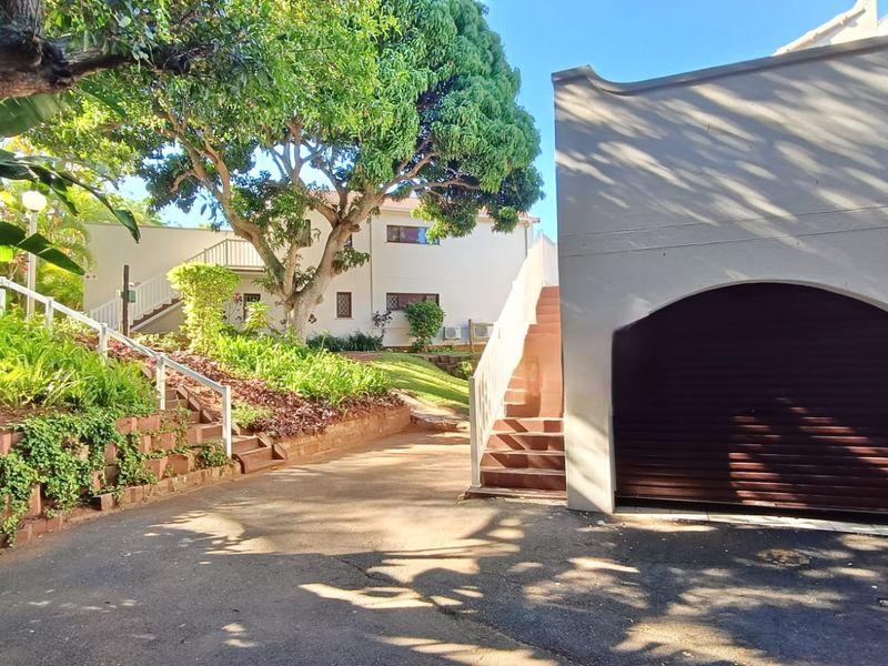 Exterior view of the property featuring a paved driveway, a single garage with a roller shutter door, and landscaped garden areas with retaining walls.