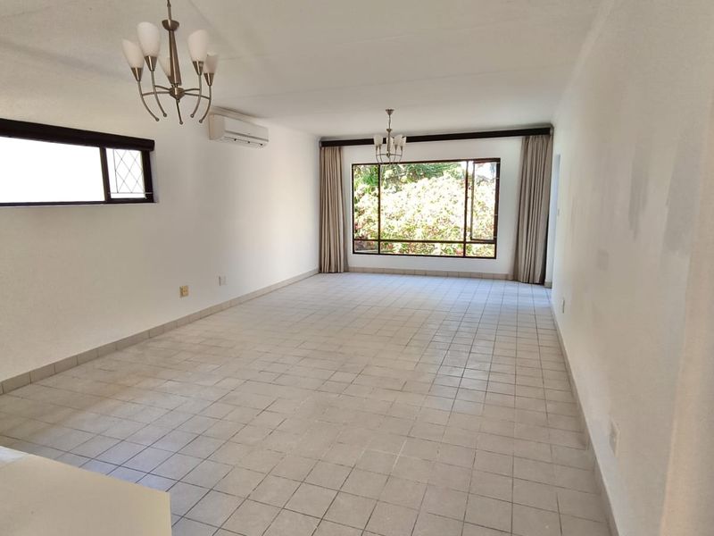 A spacious, empty living area featuring tiled flooring, a split-system air conditioner, and large windows that provide natural light and garden views.