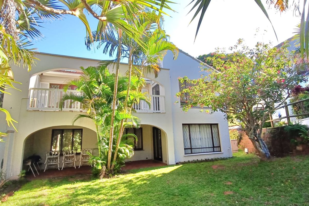 Rear view of the apartment building featuring a covered ground-floor patio, an upper-level balcony with arched detailing, and a well-maintained garden area.