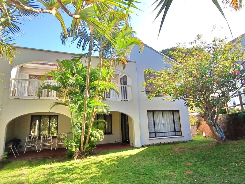 Rear view of the apartment building featuring a covered ground-floor patio, an upper-level balcony with arched detailing, and a well-maintained garden area.
