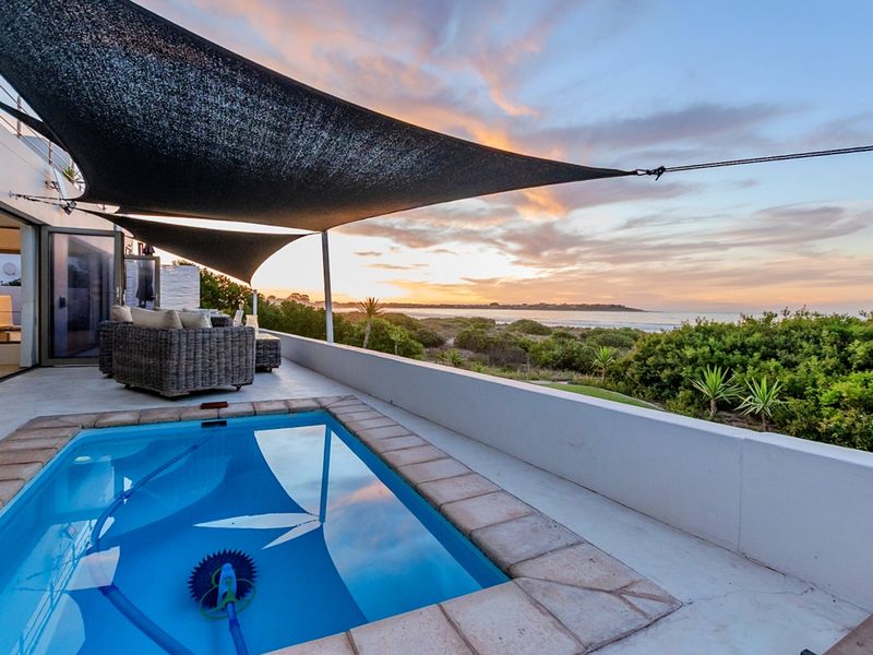 An elevated patio area featuring a swimming pool with tiled coping, a shade sail, and expansive coastal views at sunset.
