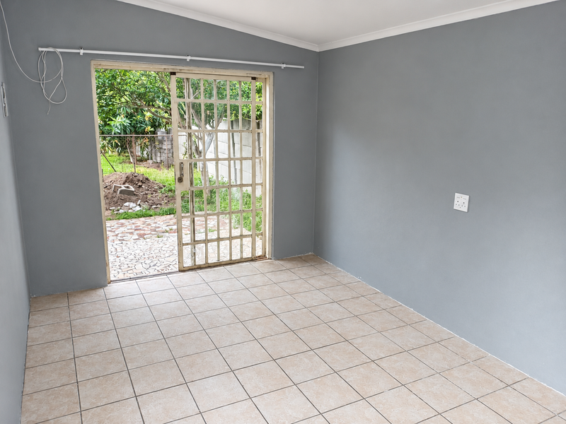 An empty living room featuring neutral grey walls, tiled flooring, and a sliding glass door with a security gate leading to the backyard.