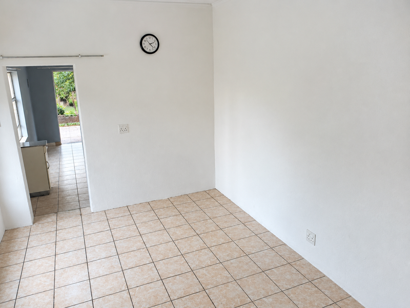 A bright, empty living area featuring neutral ceramic floor tiles and white walls, with a clear view through to an adjacent room and outdoor space.