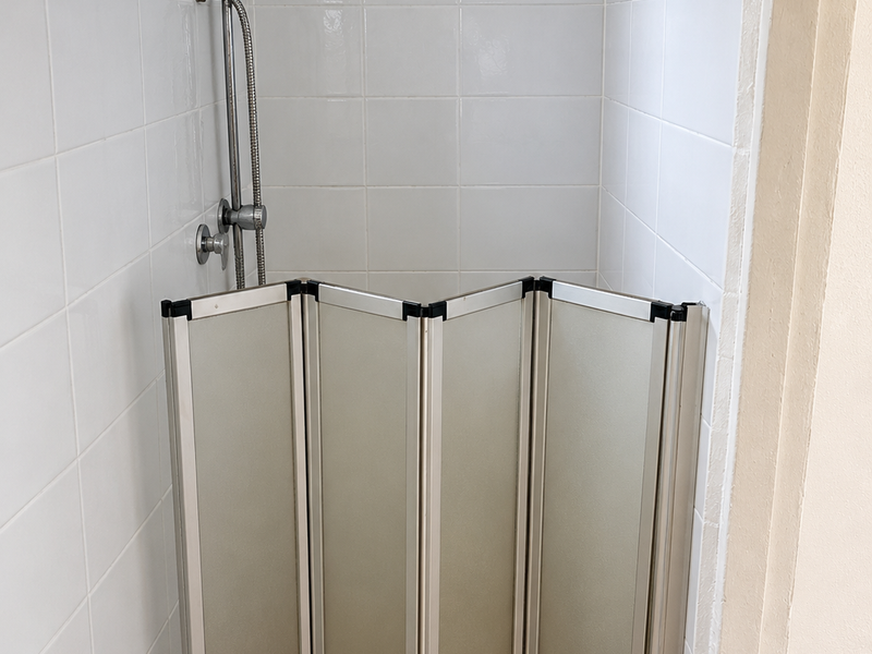 A compact shower stall featuring white wall tiles and a folding shower screen, requiring modernization of fixtures and finishes.