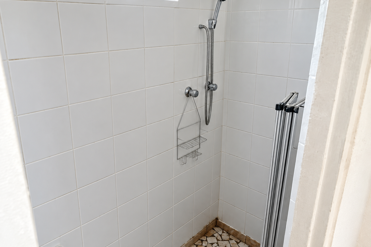 A compact shower enclosure featuring white wall tiles, a handheld shower fixture, and a tiled floor, requiring some maintenance and cosmetic updates.