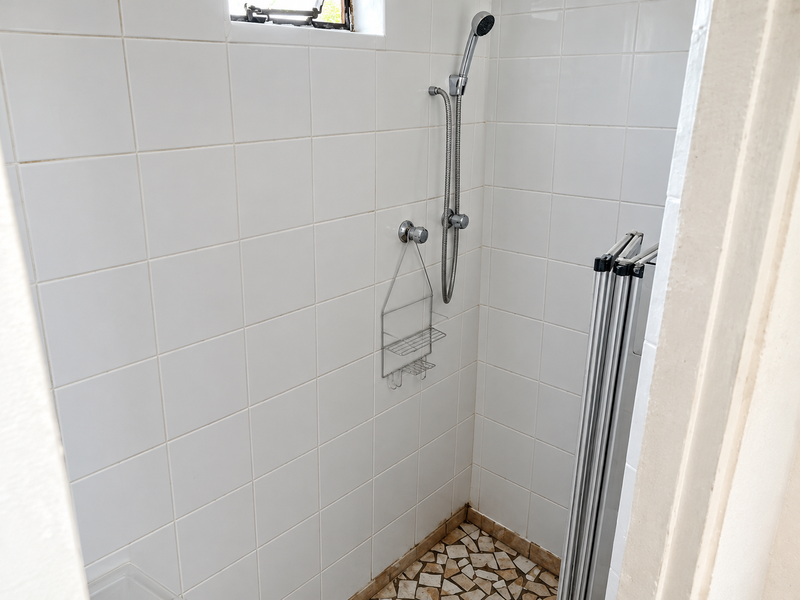 A compact shower enclosure featuring white wall tiles, a handheld shower fixture, and a tiled floor, requiring some maintenance and cosmetic updates.