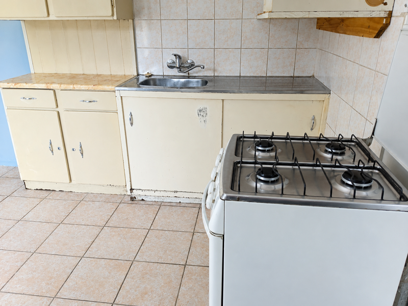 A functional kitchen featuring dated cabinetry, a stainless steel sink, tiled splashbacks, and a freestanding gas stove with ceramic floor tiles.