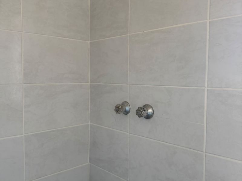 A functional shower enclosure featuring light grey ceramic wall tiles, a standard shower head, and chrome mixer taps.