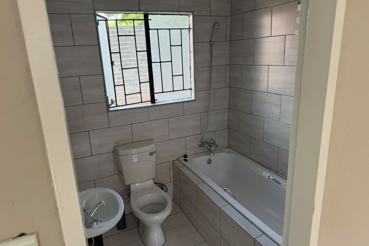 A clean, modern bathroom featuring light grey wall tiles, a white bathtub with a handheld shower mixer, a pedestal basin, and a toilet.