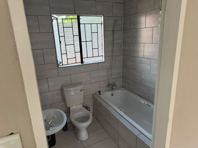 A clean, modern bathroom featuring light grey wall tiles, a white bathtub with a handheld shower mixer, a pedestal basin, and a toilet.