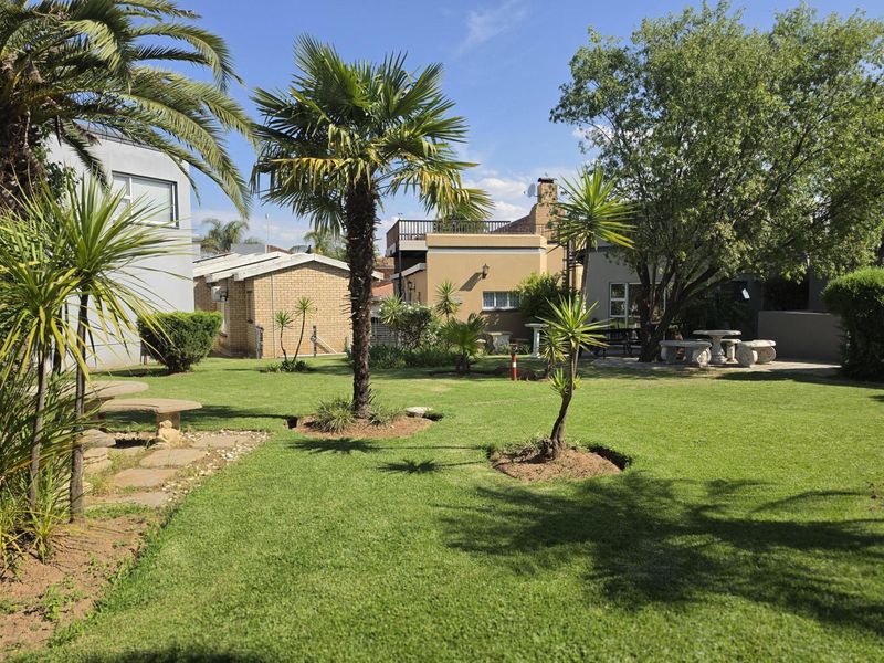 A well-maintained garden featuring a lush lawn, mature palm trees, and a paved patio area, set against a backdrop of brick residential structures.