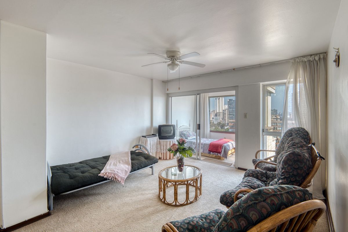 A living room area featuring carpet flooring, a ceiling fan, and sliding glass doors that open to a balcony with city views.