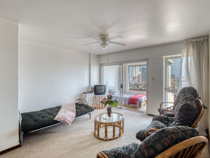 A living room area featuring carpet flooring, a ceiling fan, and sliding glass doors that open to a balcony with city views.