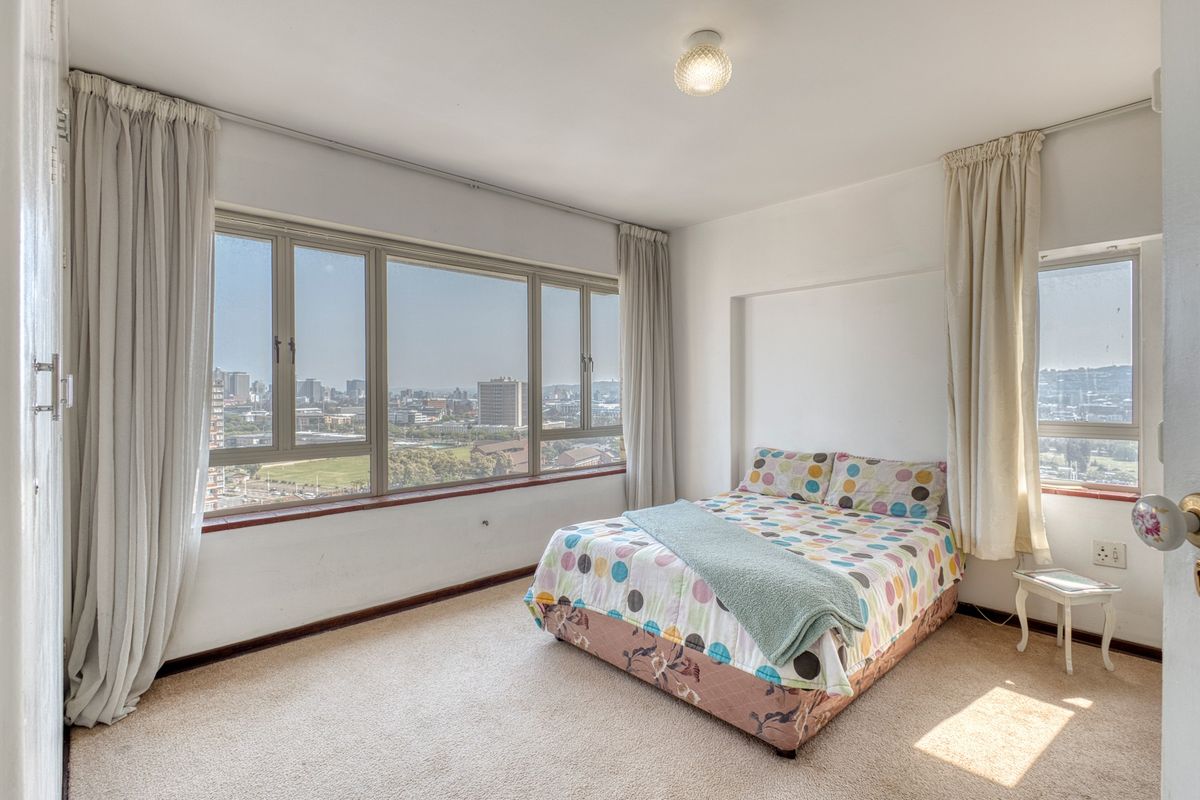 A bright bedroom featuring expansive windows with panoramic city views, neutral carpeting, and ample natural light.