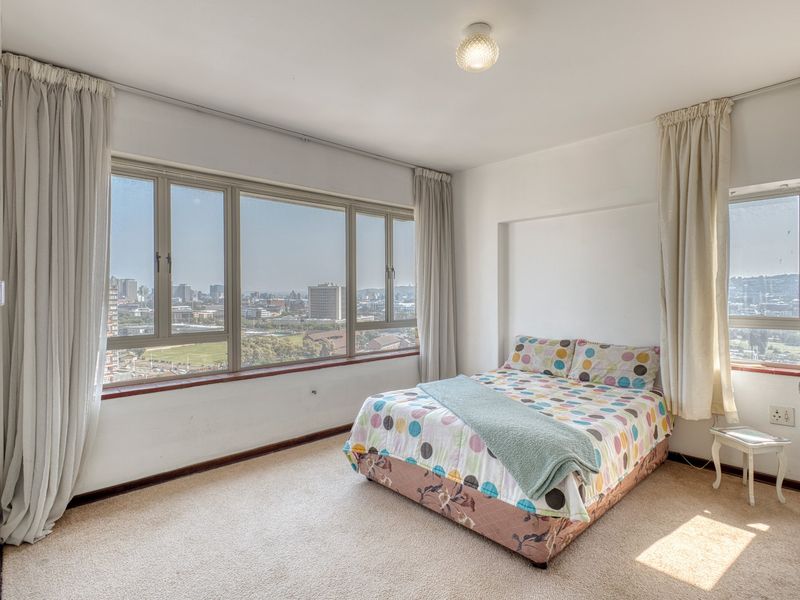 A bright bedroom featuring expansive windows with panoramic city views, neutral carpeting, and ample natural light.