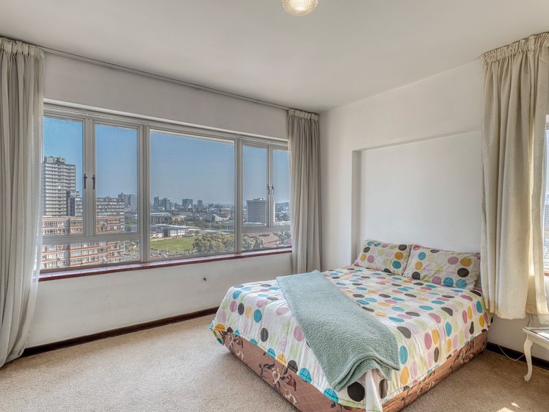 A bright bedroom featuring expansive windows with panoramic city views, neutral walls, and carpeted flooring.
