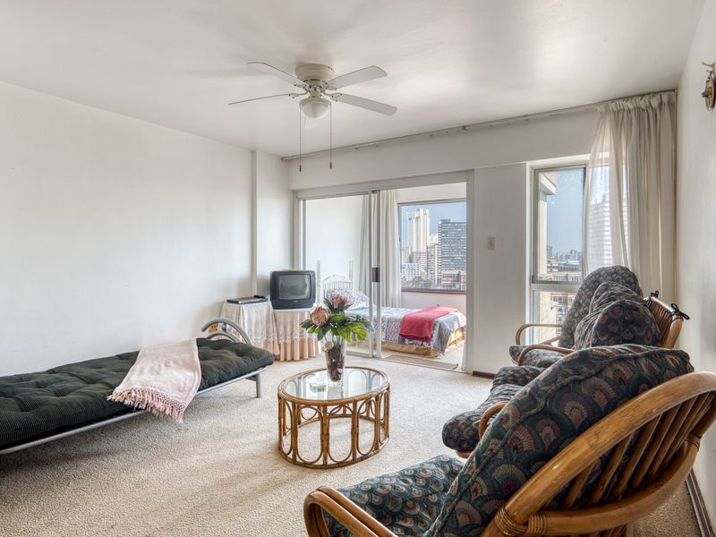 A living room featuring carpet flooring, a ceiling fan, and sliding glass doors that provide access to a balcony with city views.