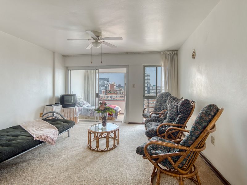 A spacious living room featuring neutral carpeting, a ceiling fan, and large sliding glass doors that open onto a balcony with city views.