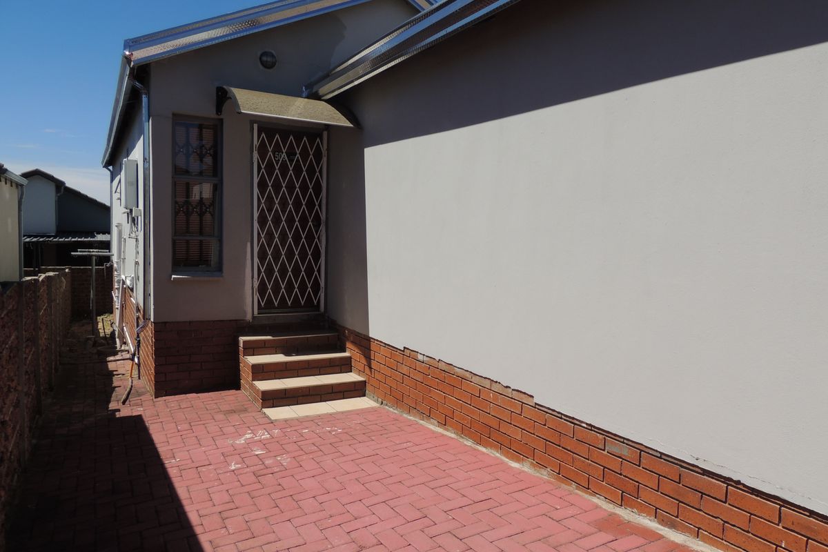 Rear exterior view showing a paved patio area, brick plinth detailing, and a secure entrance door with a protective awning.