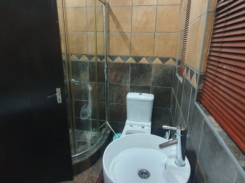 A compact bathroom featuring a corner shower enclosure, tiled walls, a modern vessel sink with chrome mixer, and wooden venetian blinds.