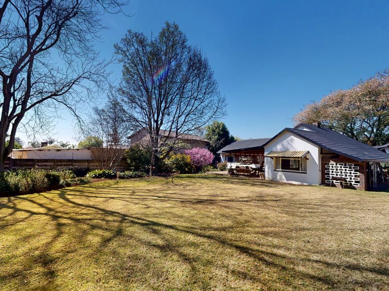 Spacious rear garden featuring a well-maintained lawn, mature trees, and a covered patio area with decorative brickwork and a tiled roof.