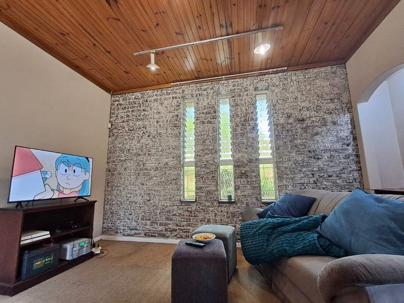 A cozy living room featuring a rustic exposed timber ceiling, a textured brick feature wall, and vertical louvre windows providing natural light.