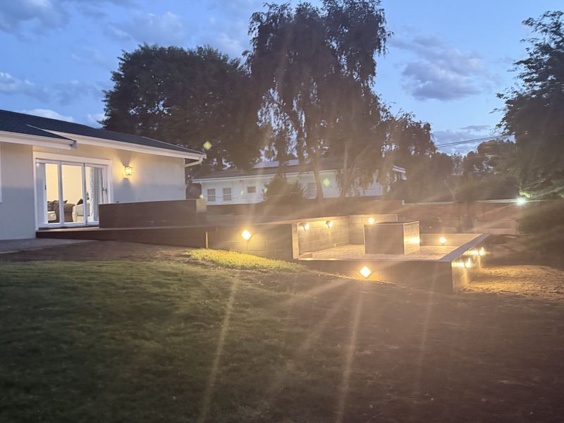 A well-lit outdoor patio area featuring a raised timber deck, a built-in fire pit, and integrated landscape lighting, perfect for evening entertaining.
