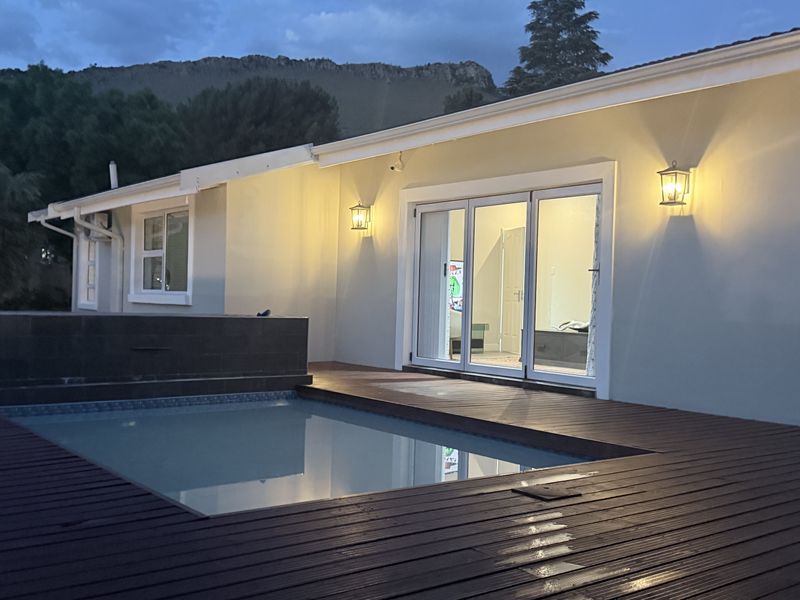 A well-maintained outdoor pool area featuring timber decking, exterior wall lighting, and sliding glass doors leading into the home at dusk.
