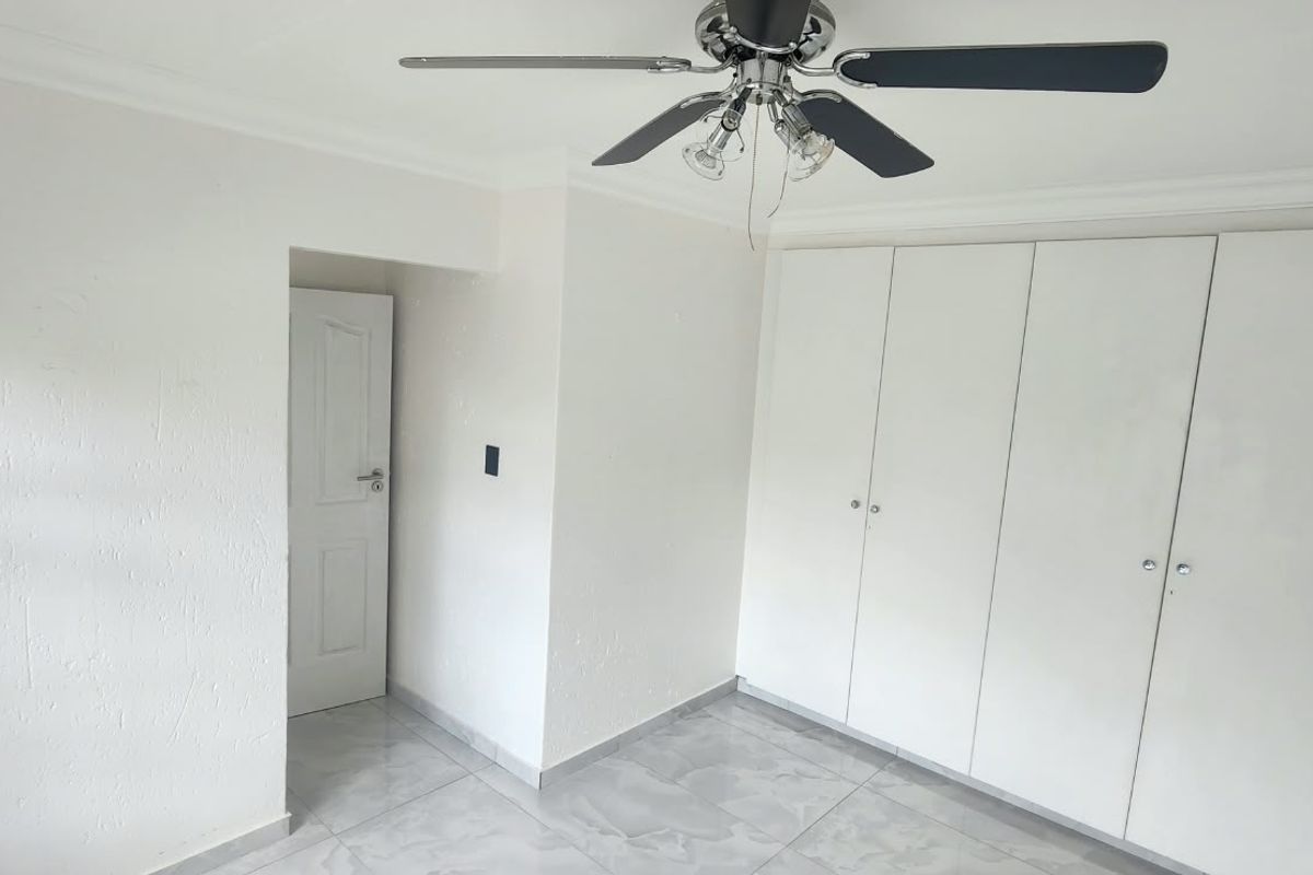 A bright, empty bedroom featuring built-in cupboards, a ceiling fan, and neutral tiled flooring.