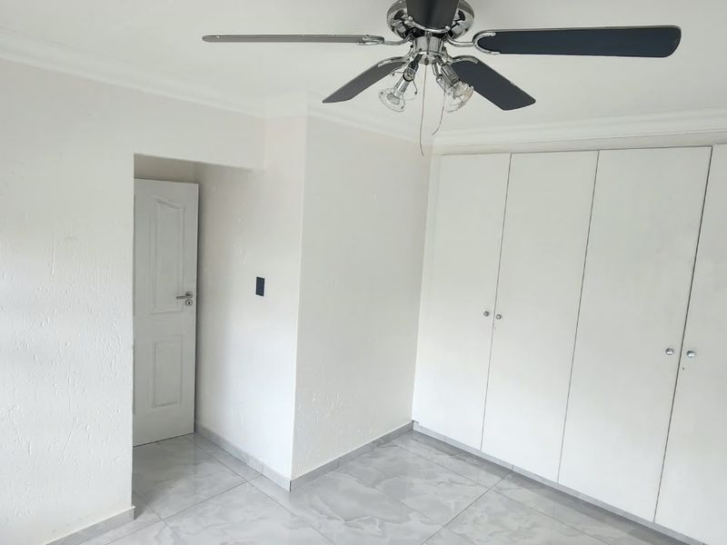 A bright, empty bedroom featuring built-in cupboards, a ceiling fan, and neutral tiled flooring.