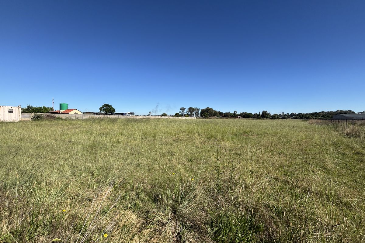 A wide-angle view of a vacant residential plot featuring open grassland, perimeter fencing, and a water storage tank under a clear blue sky.
