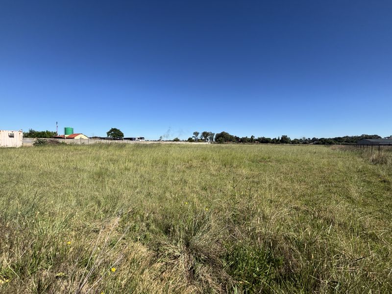 A wide-angle view of a vacant residential plot featuring open grassland, perimeter fencing, and a water storage tank under a clear blue sky.