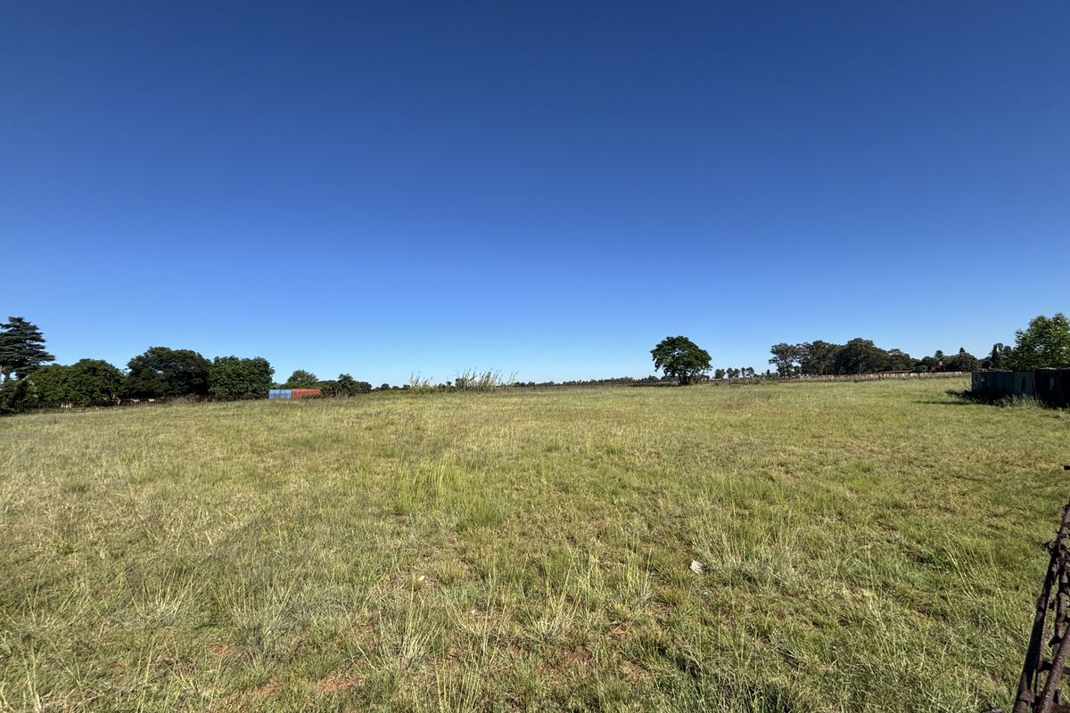 A wide, open expanse of vacant residential land featuring natural grass cover, perimeter wire fencing, and scattered mature trees under a clear blue sky.