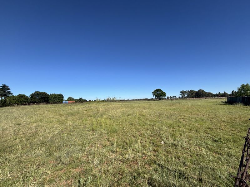 A wide, open expanse of vacant residential land featuring natural grass cover, perimeter wire fencing, and scattered mature trees under a clear blue sky.