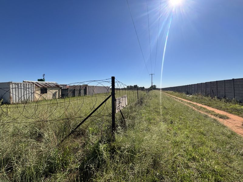 A view of the vacant residential land featuring a barbed wire fence, precast concrete boundary wall, and overhead power lines along a dirt path.