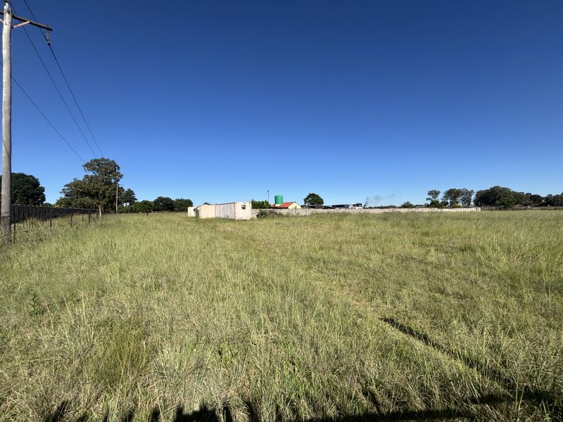 A wide-angle view of a vacant residential plot featuring open grassland, perimeter fencing, and a utility pole under a clear blue sky.