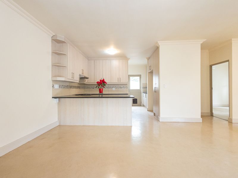 A clean, open-plan kitchen featuring light-toned cabinetry, dark stone countertops, a tiled backsplash, and integrated cooking appliances, presented in a bright, vacant space.