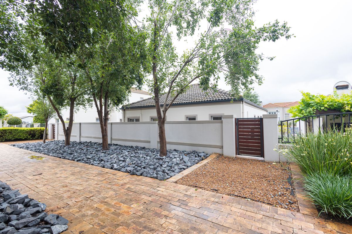 A well-maintained rear exterior area featuring a paved driveway, decorative rock garden, mature trees, and a secure timber pedestrian gate set within a boundary wall.