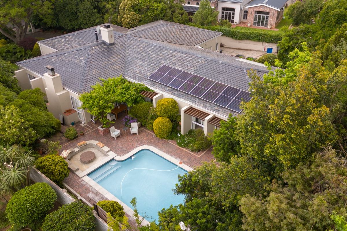 Aerial view of a residential property featuring a swimming pool, brick patio area, and roof-mounted solar panels set within a lush, landscaped garden.