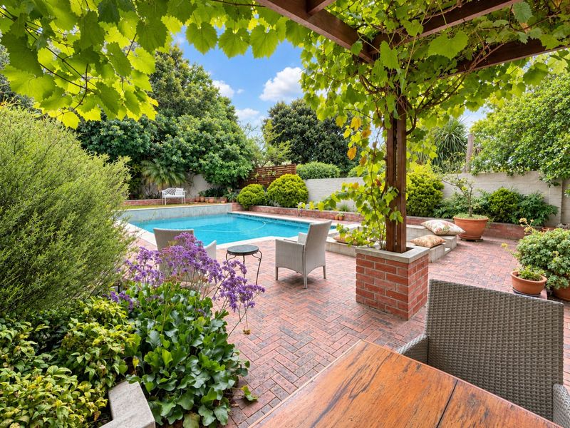 A well-maintained outdoor pool area featuring a brick-paved patio, a timber pergola with climbing vines, and established garden landscaping.