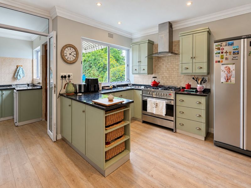 Well-appointed kitchen featuring sage green shaker cabinetry, granite countertops, stainless steel gas range, and an adjoining scullery area with light wood-look flooring.