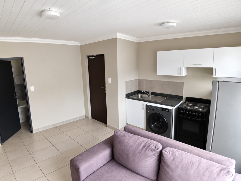 Open-plan living area featuring a compact kitchen with built-in cabinetry, stove, and integrated laundry facilities, finished with neutral tones and tiled flooring.
