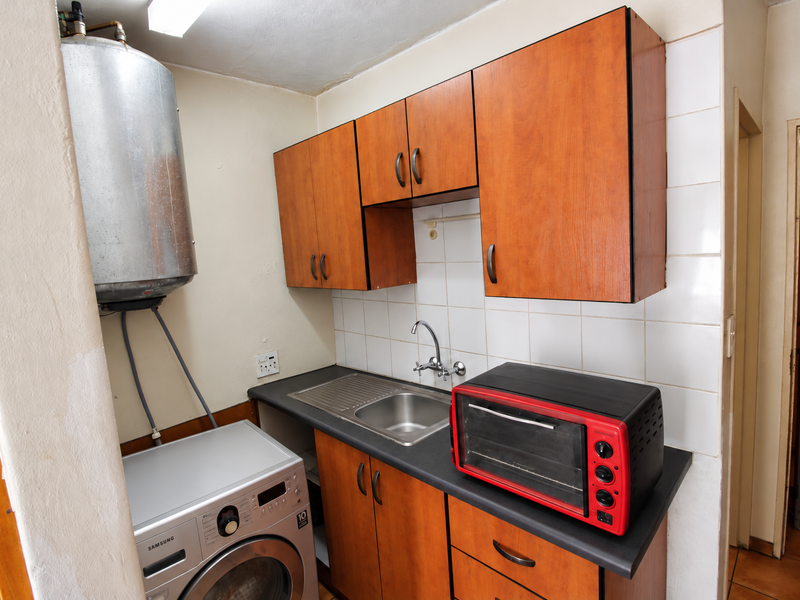 A functional laundry area featuring built-in wooden cabinetry, a stainless steel sink, tiled splashback, and an exposed electric geyser unit.