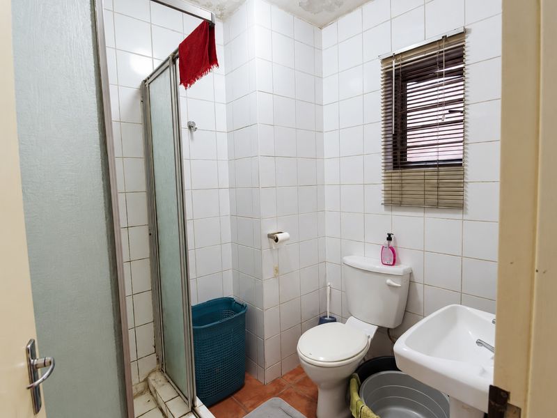 A compact bathroom featuring a shower enclosure, pedestal basin, and toilet, with white wall tiling and terracotta floor tiles. Requires cosmetic updates.