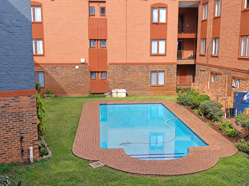 A communal swimming pool area set within a landscaped garden, surrounded by the brick facade of an apartment complex.