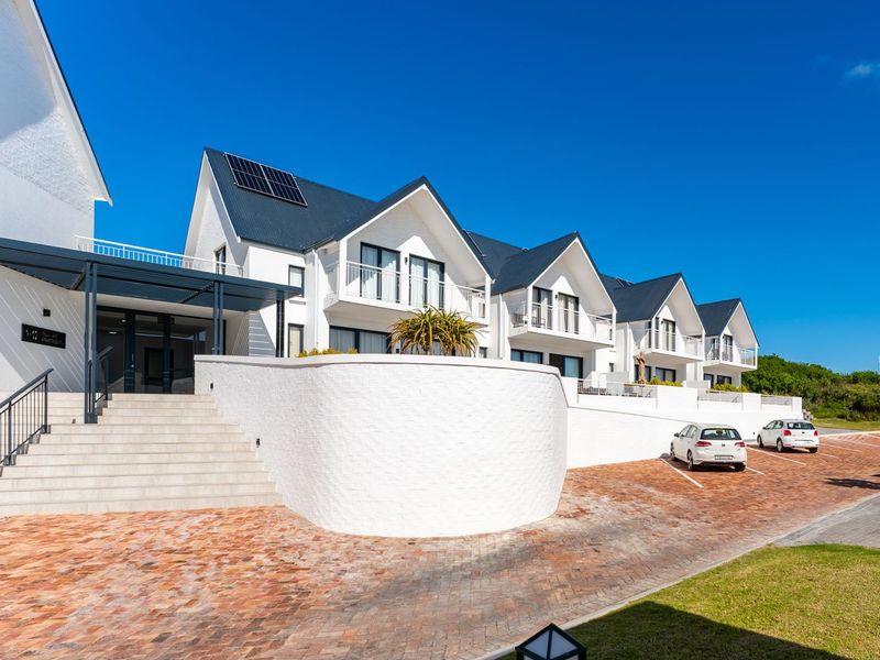 A modern townhouse complex featuring white facades, pitched roofs with solar panels, paved driveway, and elevated balconies under a clear blue sky.