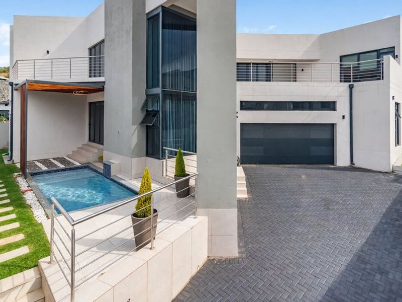 Modern rear exterior featuring a paved driveway, double garage, swimming pool, and elevated balcony with stainless steel balustrades.