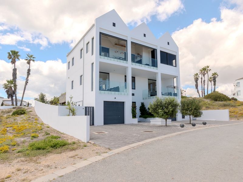 A modern three-story residence featuring a distinctive triple-gabled roofline, glass-fronted balconies, and a paved driveway with integrated garage access.