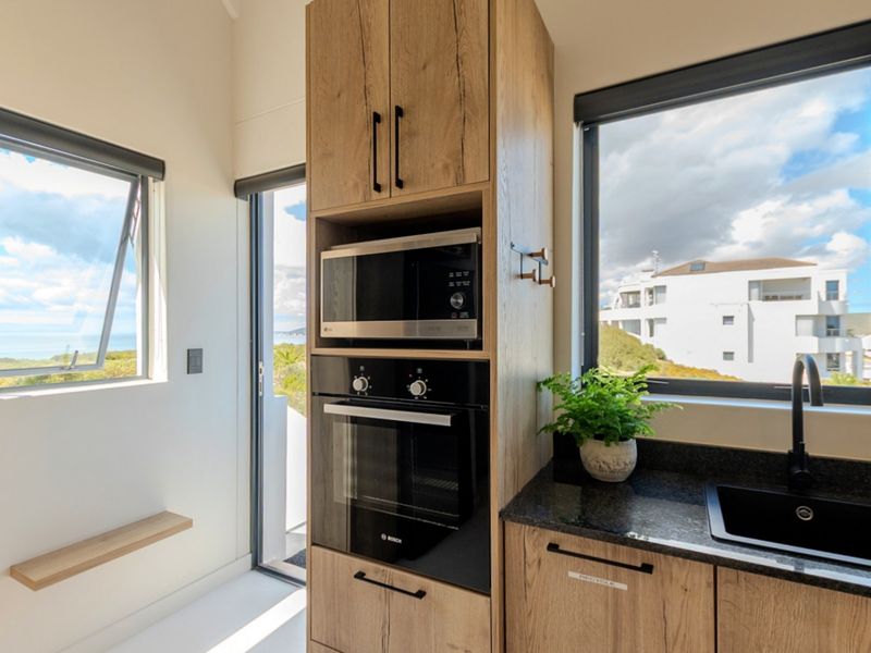 A modern, compact scullery featuring timber cabinetry, stone countertops, integrated oven and microwave, and a sleek black sink with views.