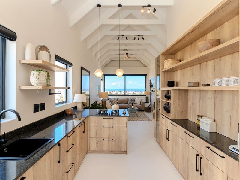 Modern open-plan kitchen and living area featuring vaulted ceilings with exposed trusses, sleek wood cabinetry, stone countertops, and expansive coastal views.